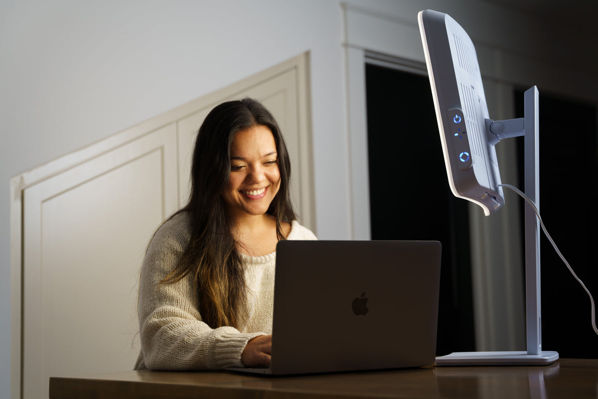 Woman smiling while working on a laptop next to the Day-Light Elite light therapy lamp