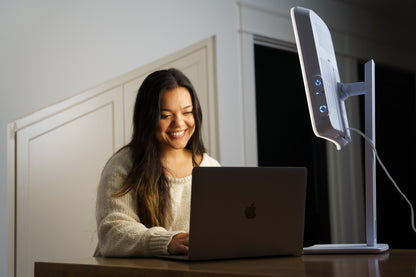 Woman smiling while working on a laptop next to the Day-Light Elite light therapy lamp