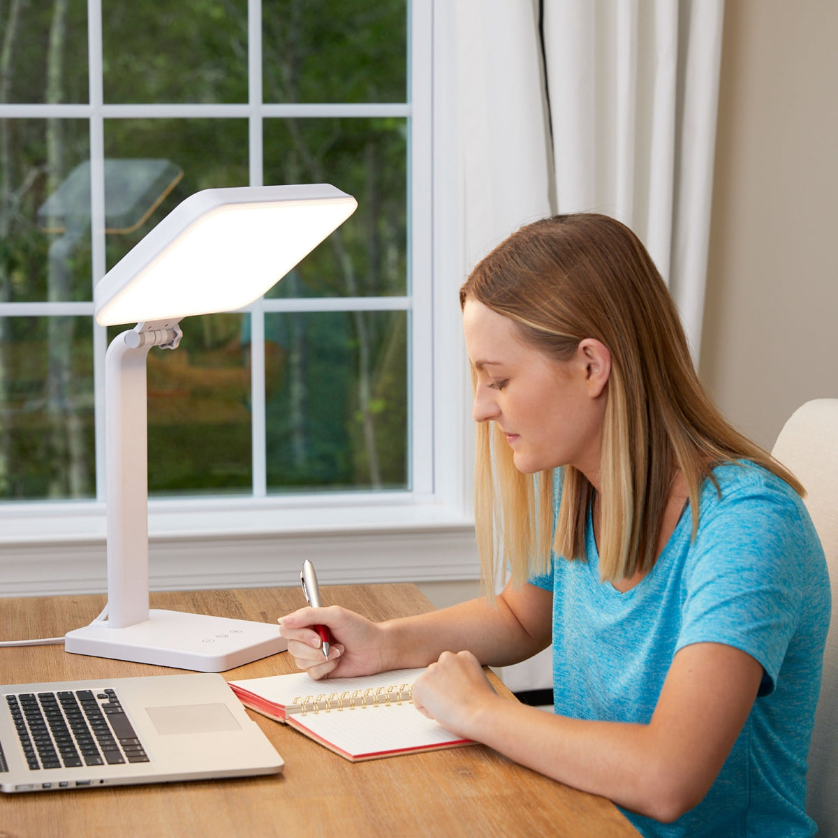 Woman writing at a home office desk under the TheraLite Aura light therapy lamp for office use